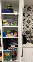 Shelf view of various toys including container of multiple Hot Wheels cars, wooden toy tool set, bagged toys and craft markers on shelf.