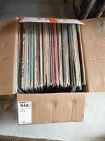 Open cardboard box filled with a variety of vertically stacked LP vinyl records showing numerous different album cover edges.
