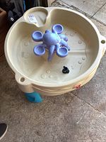 Top and angled views of beige plastic water table with blue legs. Central purple octopus water spinner. Worn surface.