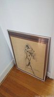 Framed unsigned sketch art shown standing against wall on hardwood floor, view shows full framed art with brown mat and figure sketch.