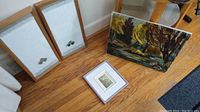 Framed canvas painting, two framed towels, and small framed artwork arranged on hardwood floor near walls