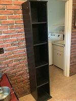 Tall black narrow bookcase standing between a brick wall and doorway, showing four shelves and visible scuff marks.