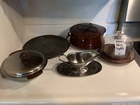 Front view of the Dansk enamelled metal casserole, metal gravy boat with tray, glass and teak cheese server, vintage hammered metal tray, and an additional metal serving dish on a shelf.
