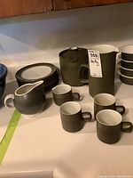Set of Denby pottery including a teapot with lid, pitcher, three mugs, creamer, nine salad plates, and a Pfaltzgraff jug on countertop.