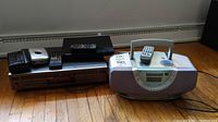 Panasonic portable stereo CD system, JVC compact disc player, Sony Blu-ray/DVD player, and their remotes shown on a hardwood floor near a baseboard heater.