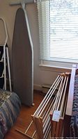 Photo shows a folded grey covered ironing board standing vertically next to two wooden clothes drying racks folded on the floor.