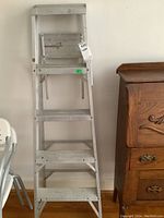 Full view of the 5-step aluminum ladder standing upright against a wall near chairs and a wooden cabinet.