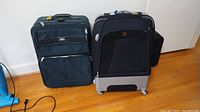 Two suitcases side by side on a wood floor: black/grey Samsonite with hard bottom and damage, dark green Heys suitcase with fabric exterior and zippered pockets