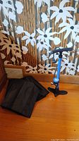 Blue Park Tool bike tire pump with black hose and pressure gauge placed on wooden floor next to black fabric hanging organizer, brown floral wallpaper background.