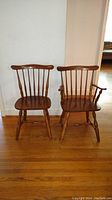 Two colonial style maple chairs pictured side by side on wooden floor. One chair has armrests and other does not.