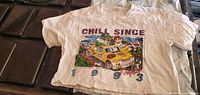 Photo of a white womens crop top showing the front graphic print with text 'CHILL SINCE 1993' and cartoon characters in a yellow car with outdoor background.