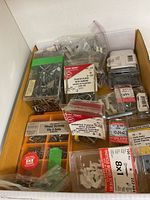 Photo of assorted screws, plastic plugs, wall anchors, and fasteners in packages and bags inside a cardboard box.