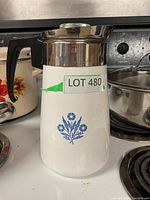 Front view of white Corning Ware coffee pot with blue flower design, stainless steel top, black handle, labeled LOT 480.