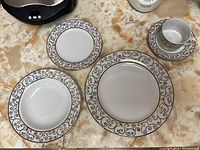 Four pieces displayed including dinner plate, salad plate, bread plate, cup, and saucer all with brown scroll pattern on white porcelain.
