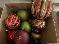 Box with assorted large Christmas balls in red, gold, green, and multicolor finishes, including several spherical ornaments with striped texture and one green glittery ball.