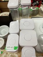 Seven OXO food storage containers with white lids displayed on a surface, showing variation in size and shape