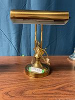 Full view of 13-inch tall brass finish piano lamp with adjustable stem and clear tubing detail around the adjustment area, positioned on wooden surface against blue curtain background.