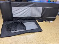 Photo showing portable massage table folded with black and gray padding and a black padded bolster on top, along with a power adapter and cords placed on the table surface.