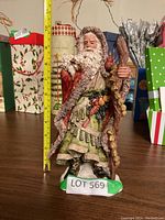 Santa Claus figurine standing on a table with measuring tape showing about 12 inches tall, wearing detailed fur-trimmed robe and holding staff.