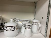 Photo showing the porcelain covered sugar bowl, creamer pitcher, salt and pepper shakers, and gravy boat, all decorated with a black scroll and gold band pattern on white porcelain.