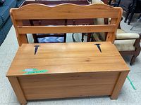 Front view of a solid wood bench showing slatted backrest and hinged seat with visible black iron hinges, natural wood finish.