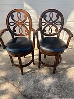 Two swivel bar stools with black leather round seats and dark wood frames, decorative backrests, and armrests, photographed outdoors in natural light.
