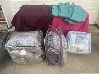 Curtain panels and comforter sets displayed on a surface; burgundy and beige curtain panels rolled up, teal grommet curtains folded, bedding sets in clear plastic packaging.