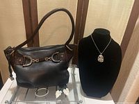 Photo shows Brighton leather hobo shoulder bag with black pebbled leather body and brown embossed leather accents along with silver-tone hardware; Brighton Adara reversible pendant displayed on black bust stand; white mother of pearl leaf-shaped dangle earrings near the bag; silver-tone crystal hinged bangle bracelet next to the bag.