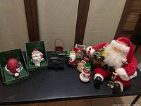 Full view of Christmas decorative items on black surface showing two stocking holders, ceramic Santa, black metal train, two glass snowmen, and plush Santa