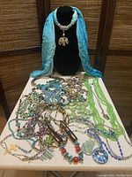 Lot of 25 mixed blue-tone necklaces laid out on a table with a light blue scarf necklace centerpiece on a jewelry bust. Variety of bead shapes, sizes, and colors, mostly blue and green hues, visible.