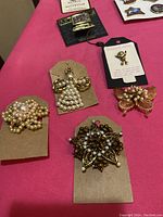 Photo showing five gold-tone brooches with pearl and bead decoration in various shapes including cluster, bow, insect, floral, and ribbon