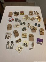 Top view photo showing 26 assorted pairs of clip-on earrings arranged on a white surface, including various shapes, colors and materials.