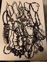 Overview photo showing tangled group of multiple black jewelry necklaces and a scarf on a cream surface.