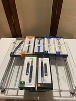 Seven Cross brand pens in original gift boxes on a metal tray. Pens include stylus, ballpoint, gel ink, and fountain styles in blue, silver, and white barrels.