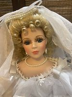 Close-up of porcelain doll head showing blonde curled hair, veil, pearl jewelry, and detailed facial features.