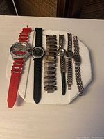Six Geneva watches placed on a white pad showing variety of bands including red silicone, black rubber, gold tone wide band with crystals, silver tone half band, and two square-faced quartz watches in metal bands showing wear.