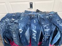 Photo of a stack of multiple ladies blue jeans showing various levels of distress, ripped holes, and paint splatter designs in different blue shades.