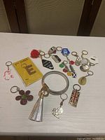 Photo showing 15 assorted key chains on white table including silver bangle wristlet, glitter letter E, London flag, pink puff, heart crystal, mother of pearl, and other metal/plastic key chains.