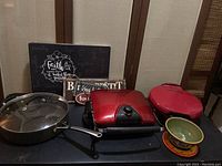 Photo shows large stainless steel skillet with glass lid, two red George Foreman grills, decorative wall hanging, and two ceramic bowls on dark surface with wood paneled background.