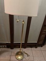 Full view of a brass swing arm floor lamp with white drum shade standing on tiled floor.