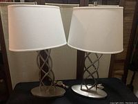 Two silver table lamps with twisted metal double-helix design and white drum shades on oval bases shown together.