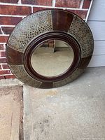 Full view of the large round mirror resting against a brick wall and concrete floor. Shows frame pattern and overall size shape.