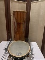 West African style talking drum with wooden body and hourglass shape positioned behind the tambourine on a glass surface