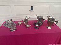 Full view of all items on a red surface showing silver plated water pitcher, 3-piece coffee/tea set, silver plated sugar bowl without lid, stainless steel tea pot, and angel figure.