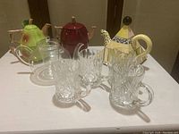 Full set photograph showing two ceramic fruit teapots (pear and apple), clear glass teapot with infuser, yellow pyramid-shaped ceramic teapot, and four crystal mugs arranged on a white surface.