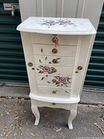 Front view of white hand painted jewelry armoire with floral designs on drawers and top, showing brass ring pulls and locking key attached.