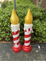 Photo showing two outdoor Christmas blow mold candle sticks with red and white striped bodies, red bases, yellow flame tops, and attached lights with cords, set outdoors in front of greenery.