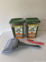 The lot items arranged on a white surface showing the litter scoop, small pink comb, and two large green containers of Greenies cat treats with value-size labels.