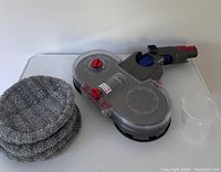 Photo showing electric mop head, six gray microfiber mop pads stacked, and clear plastic measuring cup on a white surface