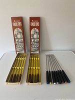 Photo showing both sets of fondue forks side by side: 8 vintage forks in yellow boxes with wooden handles and 8 modern forks with black handles and colored tips.
